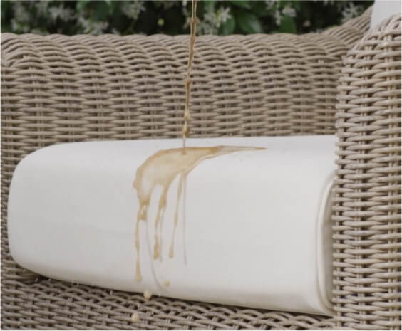 Liquid spilled on a light-colored outdoor cushion placed on a woven wicker sofa, illustrating fabric surface and stain visibility