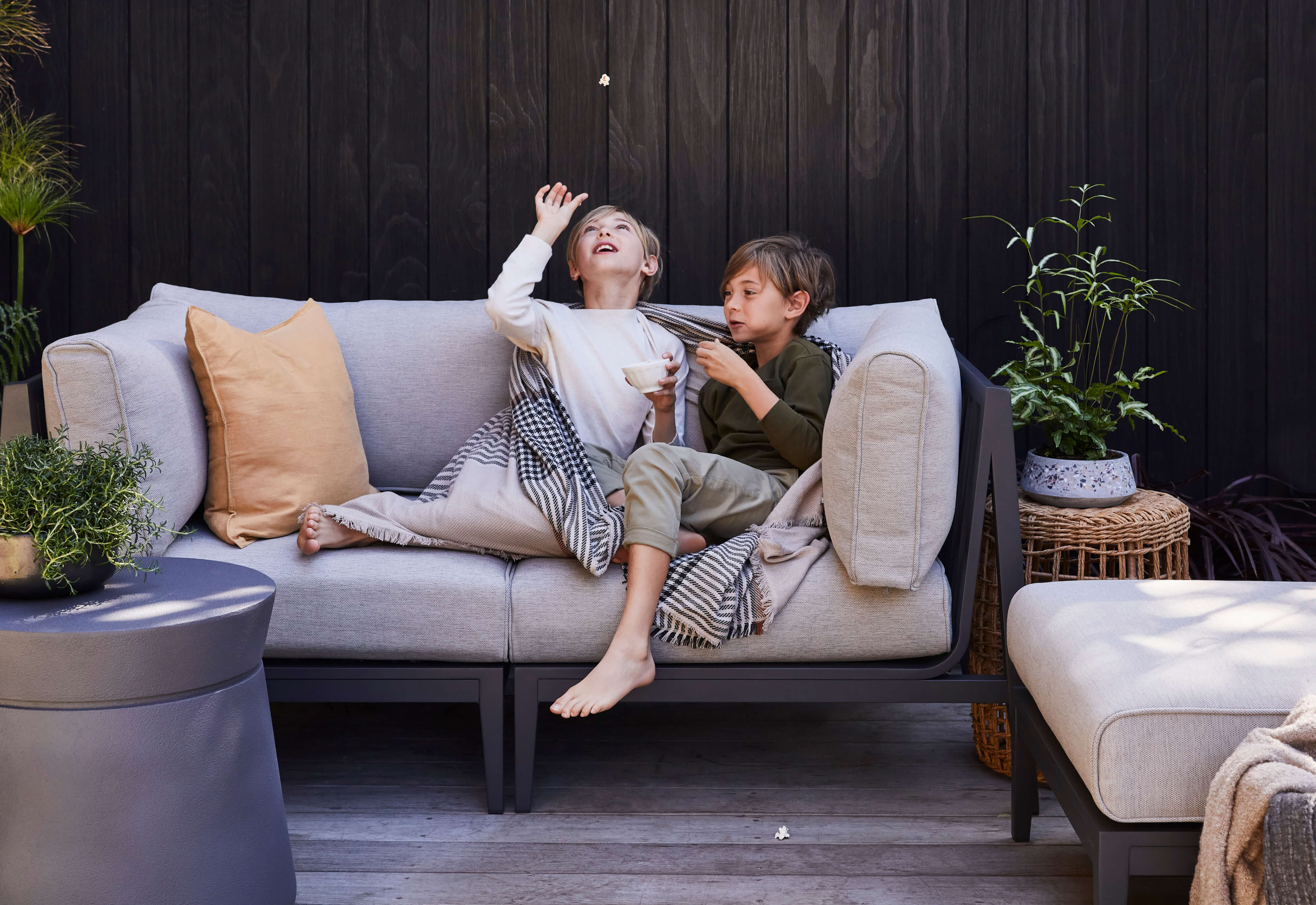 Children playing on the charcoal aluminum loveseat with sandstone gray cushions, wrapped in striped plaid bug shield blankets.