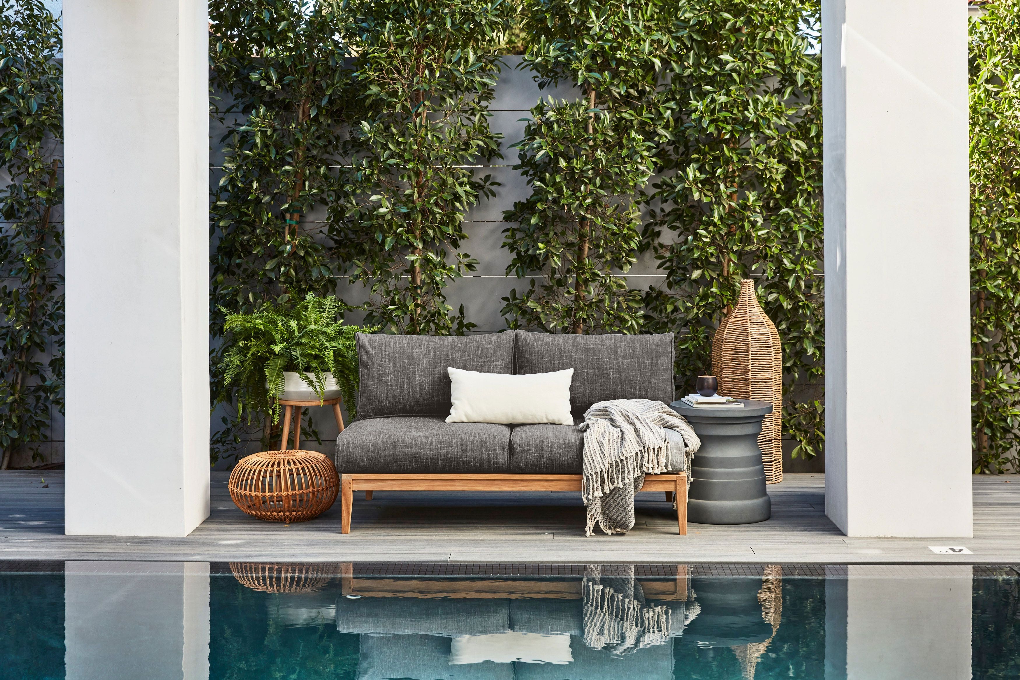 A teak outdoor sofa with matching armchairs arranged on a patio, featuring light-colored cushions and surrounded by greenery.-teak outdoor sofa-outer